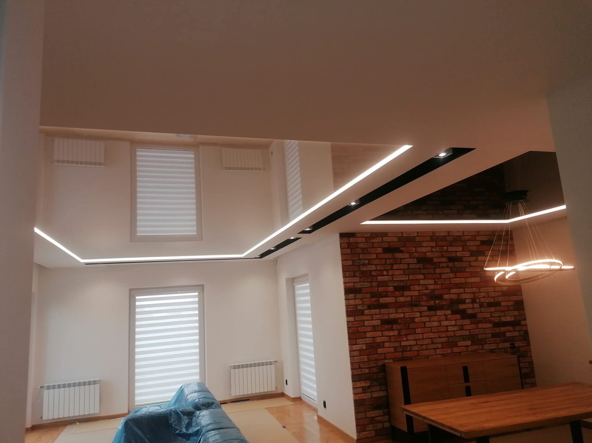 Nowoczesne wnętrze po remoncie: białe ściany, ceglana ściana akcentująca, podwieszany sufit z oświetleniem LED. Częściowo zakryte meble folią ochronną.