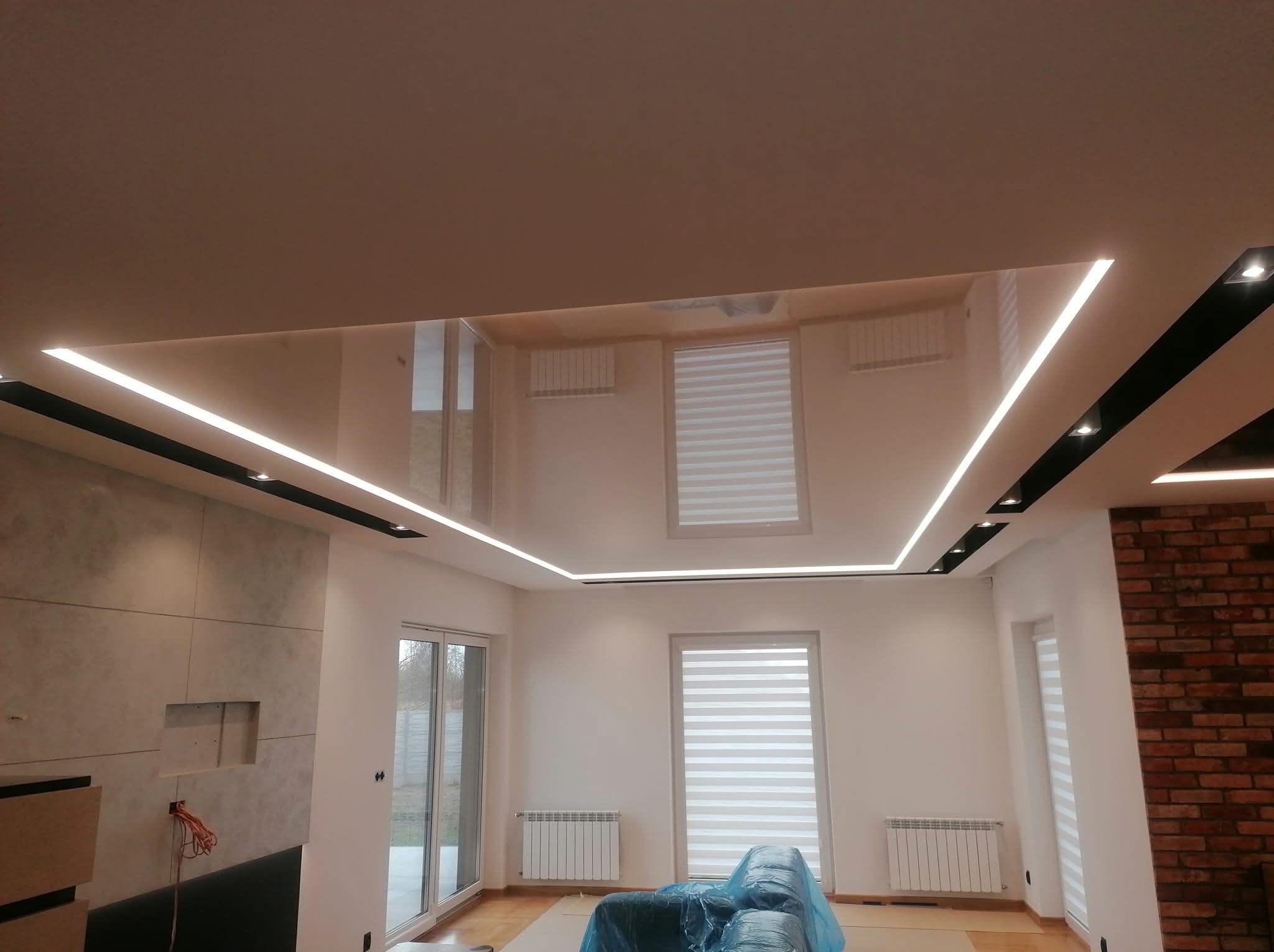 Nowoczesny salon po remoncie: podwieszany sufit z oświetleniem LED, ściana z cegły, okna z roletami. Częściowo zakryty mebel folią ochronną.