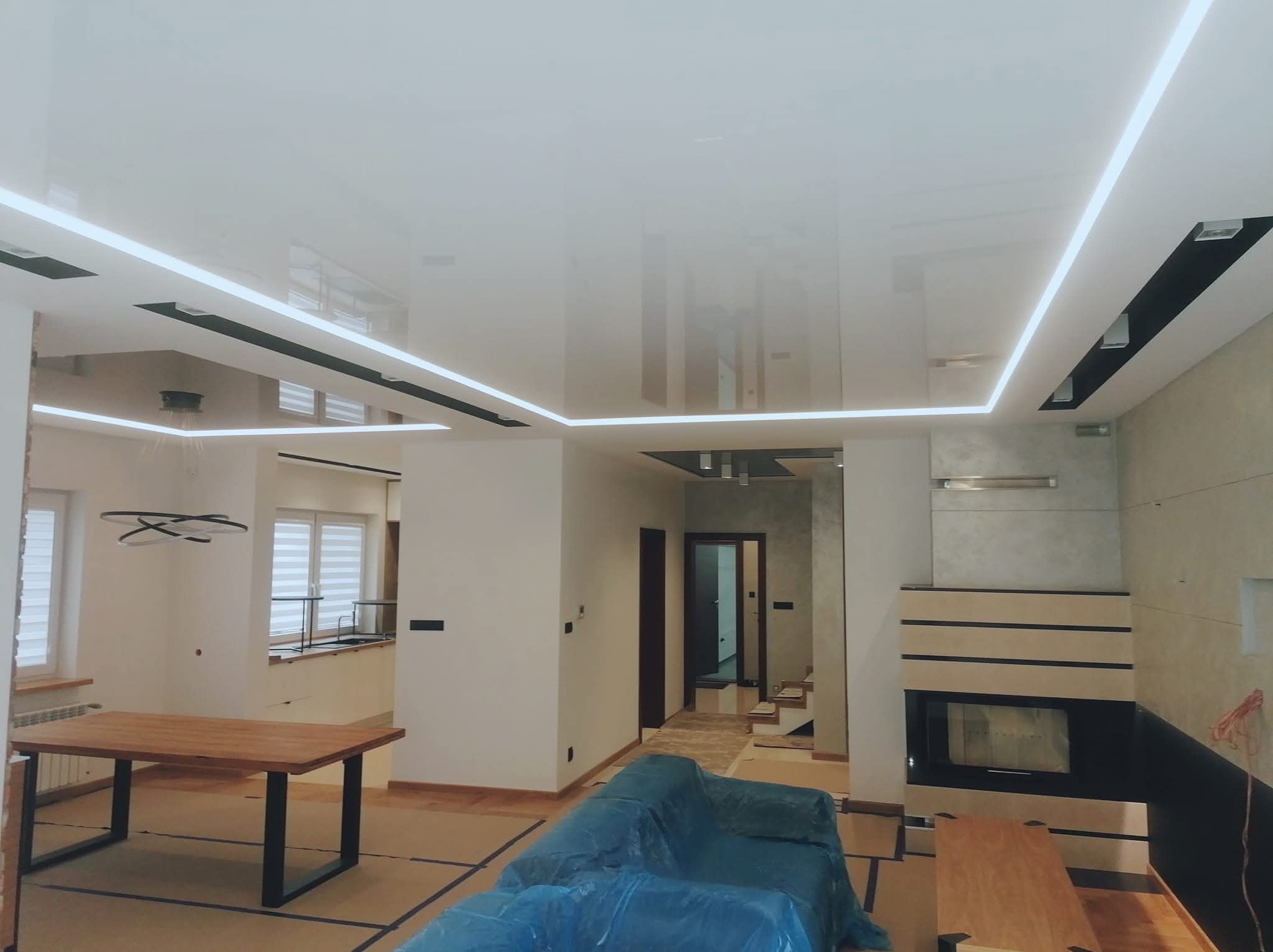 Nowoczesne wnętrze z podwieszanym, błyszczącym sufitem i liniowym oświetleniem LED. Meble zabezpieczone folią, widoczny kominek i drewniany stół.