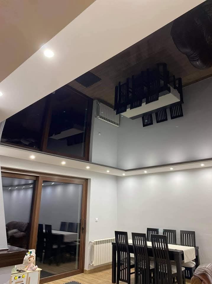 Jasne wnętrze jadalni z dużym oknem przesuwnym, szarymi ścianami i ciemnym stołem z krzesłami. Sufit podwieszany z oświetleniem punktowym. Widoczny grzejnik.