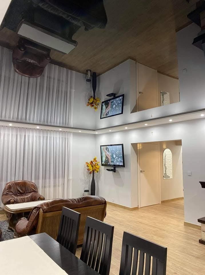 Jasne wnętrze salonu z drewnianą podłogą, białymi ścianami, telewizorami i meblami w stylu retro. Widoczne detale wykończenia i oświetlenie.