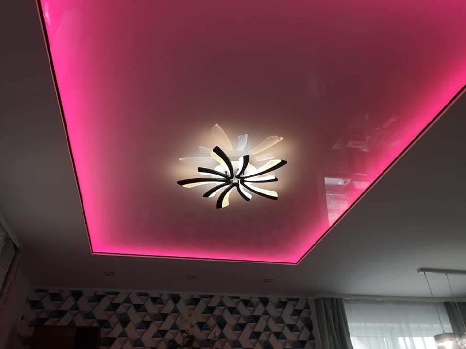Błyszczący, napinany sufit w odcieniach różu z nowoczesnym oświetleniem centralnym. Widoczny fragment wzorzystej ściany i zasłony w tle.