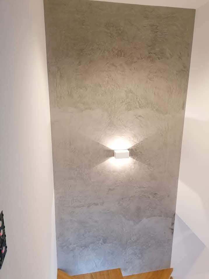 Ściana z efektem dekoracyjnym w odcieniach szarości, oświetlona nowoczesnym kinkietem. Widoczny fragment schodów i ściana boczna. Unikalna faktura gładzi.