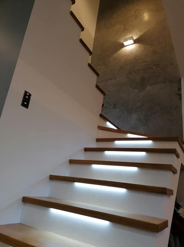 Nowoczesne schody z drewnianymi stopniami i podświetleniem LED, ściana z efektem betonu, minimalistyczna lampa ścienna. Wykończenie wnętrza w jasnych barwach.