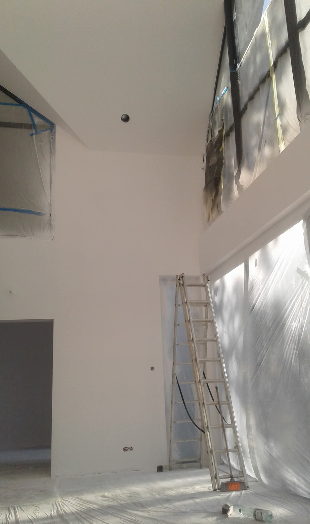 Wysokie wnętrze po malowaniu na biało, zabezpieczone folią, z drabiną opartą o ścianę. Widoczne ślady farby na folii ochronnej i podłodze.