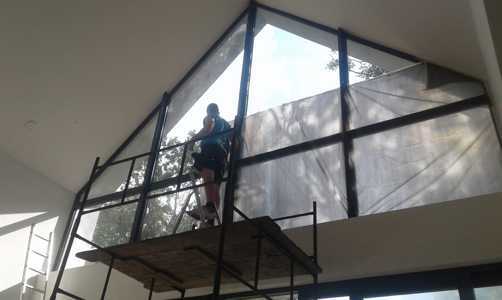 Pracownik na rusztowaniu zabezpiecza folią okno o nietypowym, trójkątnym kształcie w jasnym wnętrzu. Widoczne drzewa za oknem i drabina oparta o ścianę.