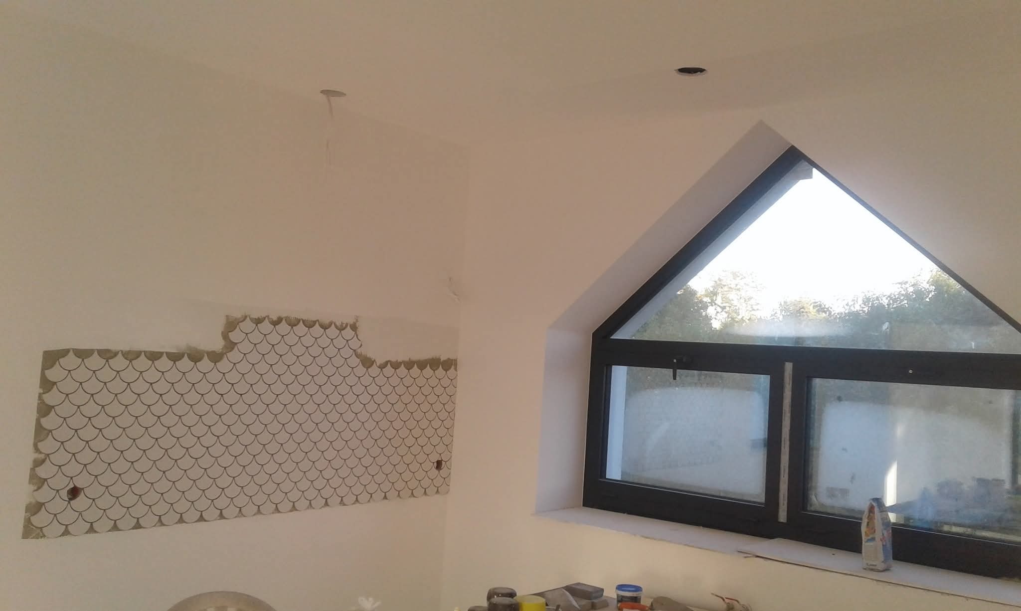 Fragment ściany z częściowo ułożoną mozaiką w kształcie rybiej łuski, widok na okno trójkątne, surowy stan wykończenia wnętrza, widoczne narzędzia i materiały budowlane.