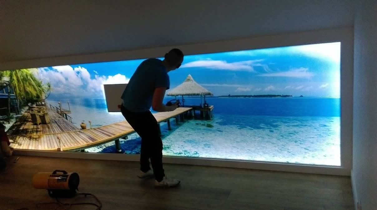 Montaż fototapety z motywem tropikalnej plaży w pomieszczeniu. Widoczny mężczyzna podczas pracy, narzędzia i fragment podłogi z panelami. Nietypowe oświetlenie.