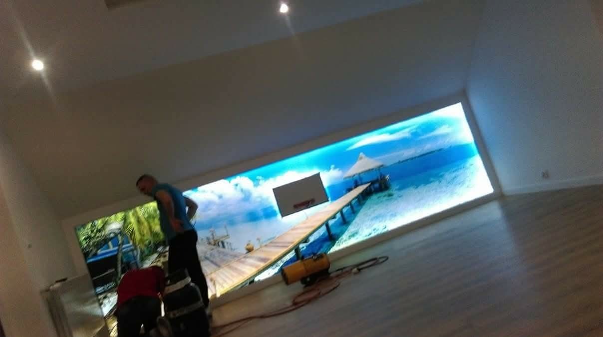 Montaż podświetlanej fototapety z motywem plaży w nowoczesnym wnętrzu. Dwóch mężczyzn przy pracy, widoczne narzędzia i fragmenty podłogi z paneli.