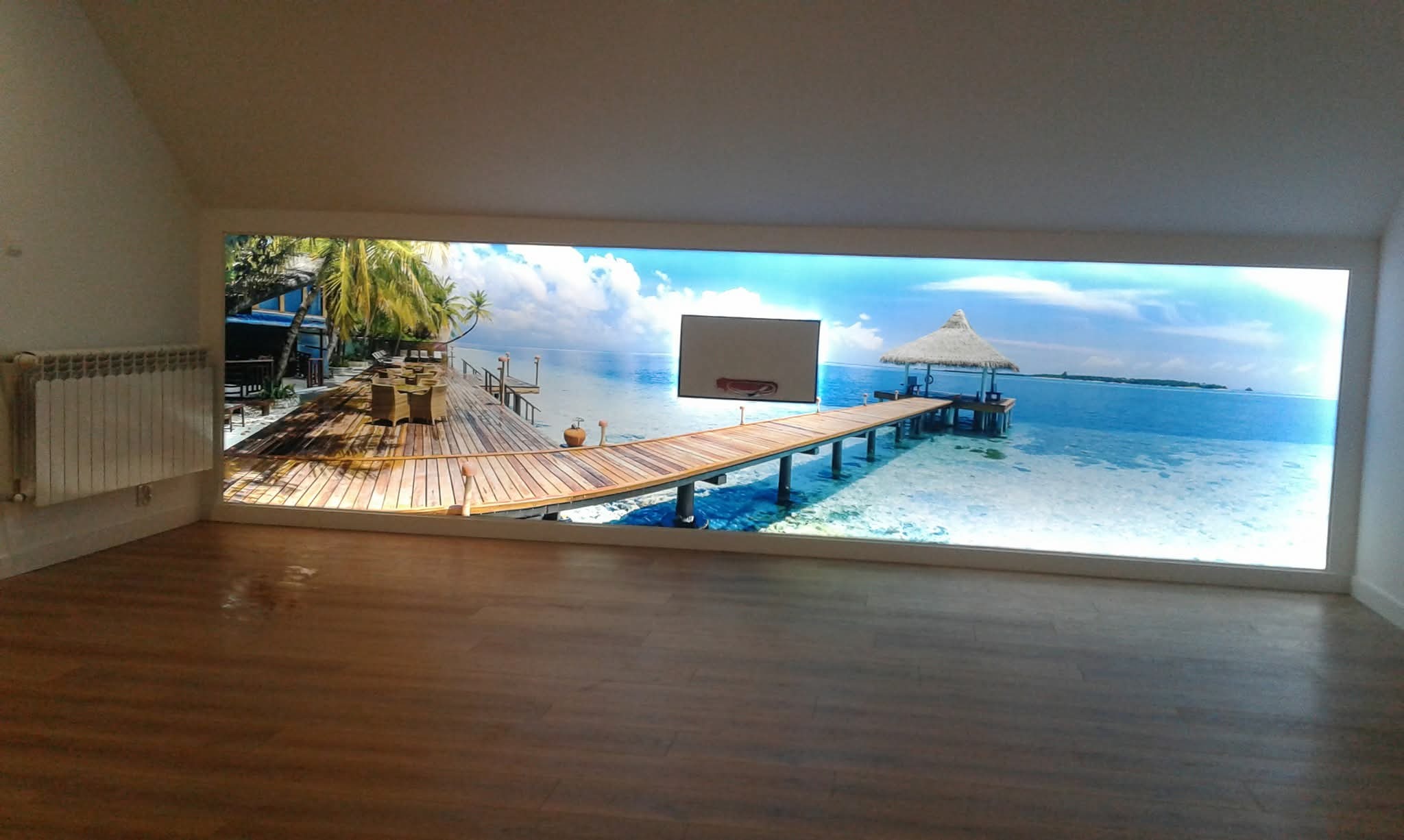 Fototapeta z motywem tropikalnej plaży na ścianie w pokoju z drewnianą podłogą. Widoczny fragment grzejnika. Nietypowe połączenie motywów.