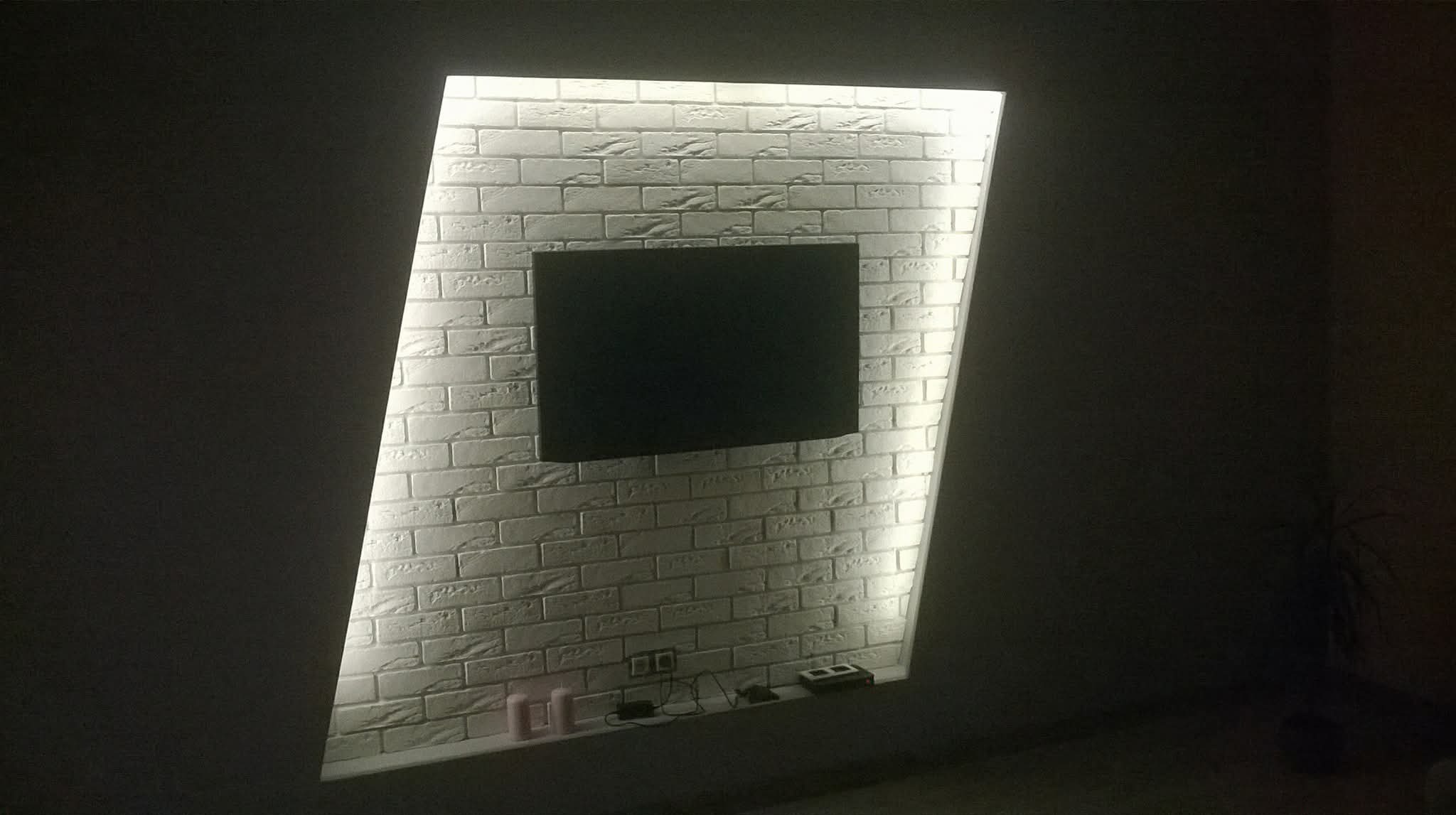 Ściana z dekoracyjną cegłą, podświetlona ledami, z zamontowanym telewizorem. Widoczne kable i dekoracje na półce. Ciemne otoczenie podkreśla efekt podświetlenia.