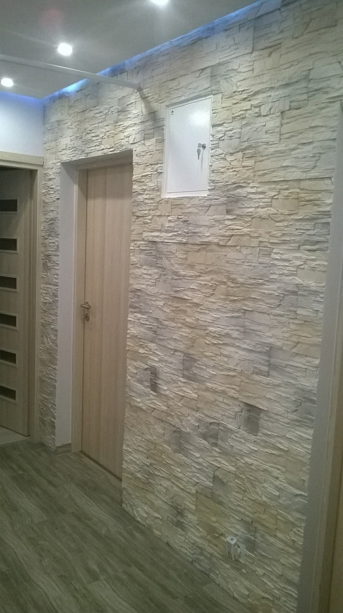 Ściana wykończona dekoracyjnym kamieniem w odcieniach beżu i szarości, z widoczną szafką elektryczną z kluczykiem. W tle fragment drzwi i podłogi imitującej drewno.
