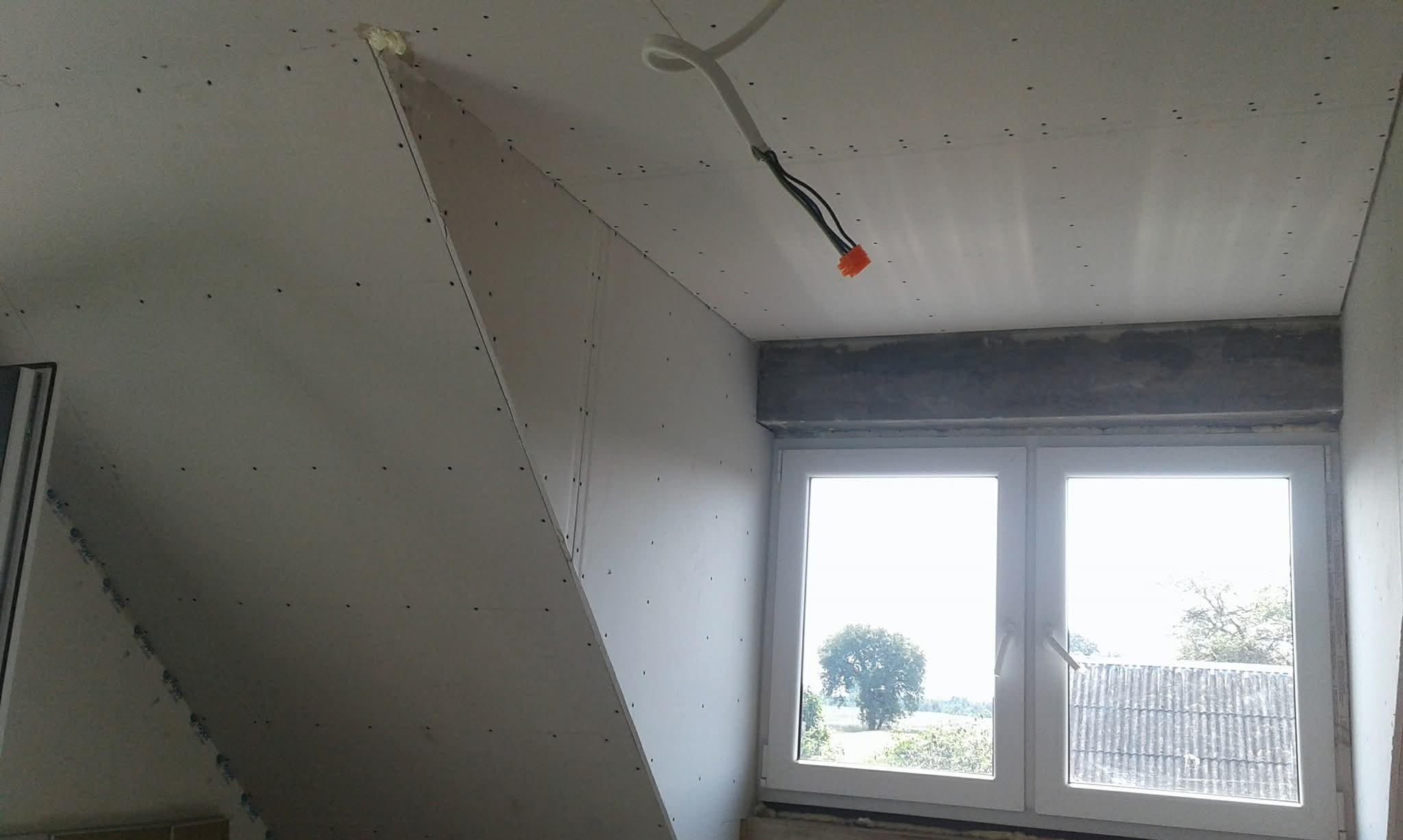 Poddasze z oknem po montażu płyt karton-gips, widoczne wkręty, przewody elektryczne. Ściana skośna. Widok na zewnątrz przez okno.
