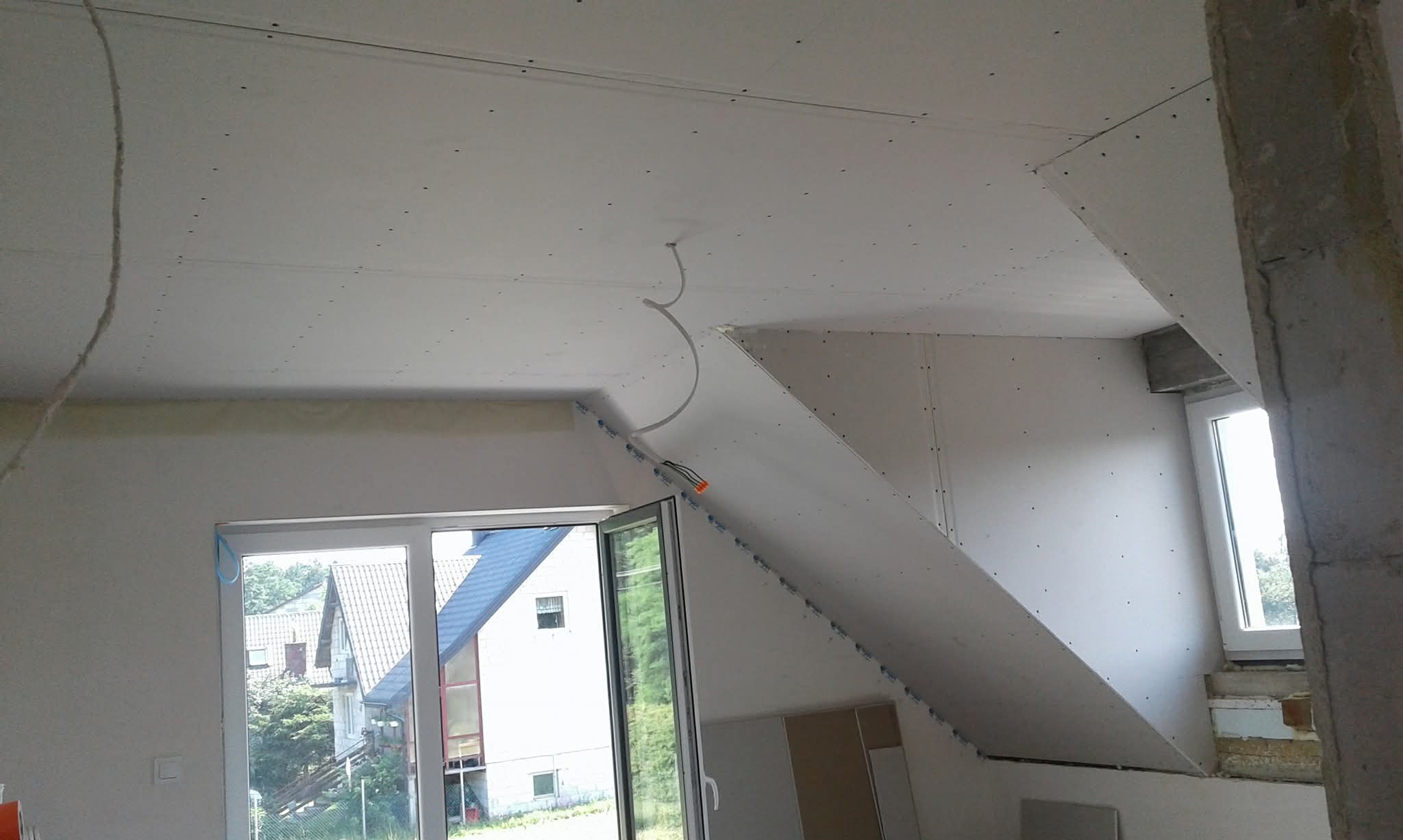 Poddasze z oknami, wykończone płytami kartonowo-gipsowymi, widoczne kable elektryczne. Prace wykończeniowe w jasnym wnętrzu, surowe wykończenie.