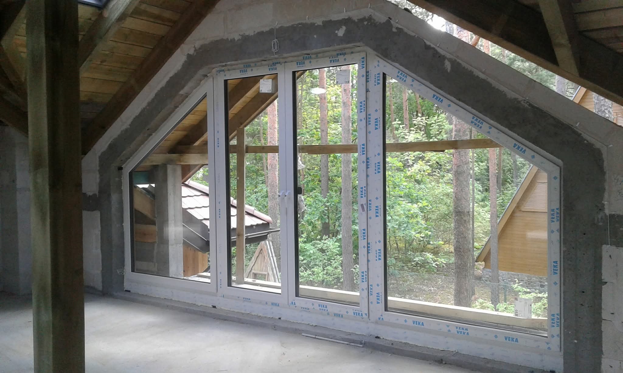 Montaż okna trzyskrzydłowego w surowym stanie poddasza. Widok na las przez okno. Białe ramy okienne z naklejkami producenta. Surowe ściany i drewniana konstrukcja dachu.