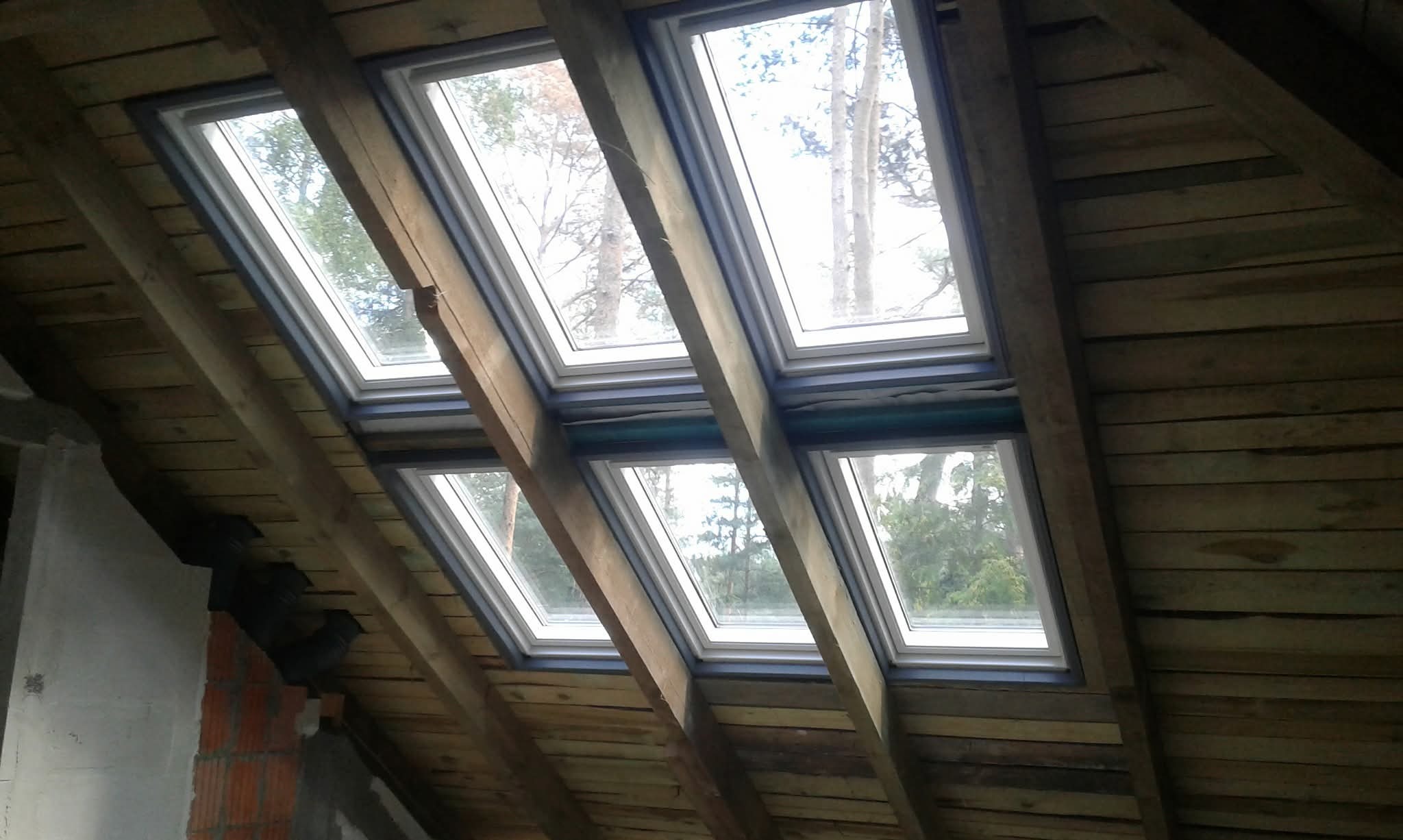 Montaż okien dachowych w drewnianej konstrukcji poddasza. Widoczne cztery okna z widokiem na zewnątrz. Częściowo otynkowana ściana z ceglanym fragmentem w lewym dolnym rogu.