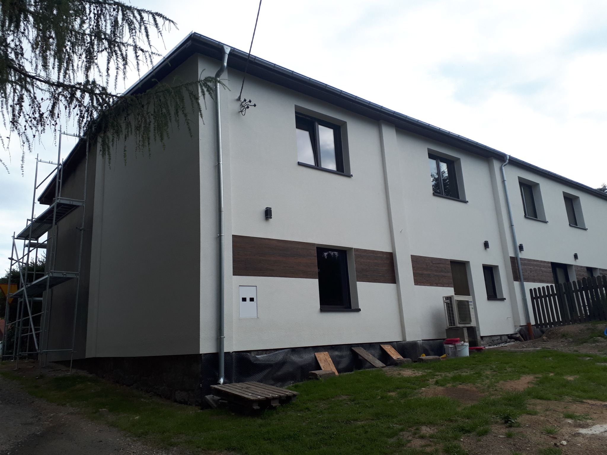 Dwukondygnacyjny budynek po renowacji elewacji, z fragmentami dekoracyjnej okładziny imitującej drewno, widoczne rusztowanie z boku budynku i elementy instalacji odprowadzającej wodę z dachu.