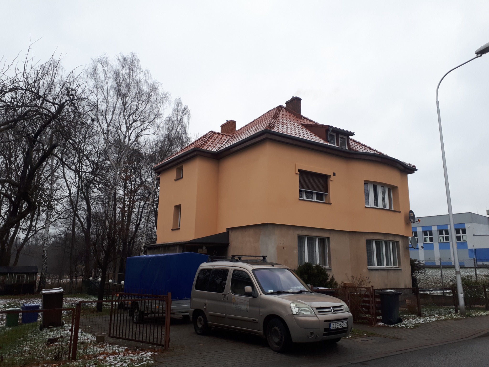 Dwupiętrowy dom z nowym, czerwonym dachem z dachówki ceramicznej, zaparkowany samochód dostawczy z przyczepą na posesji, pochmurny dzień.