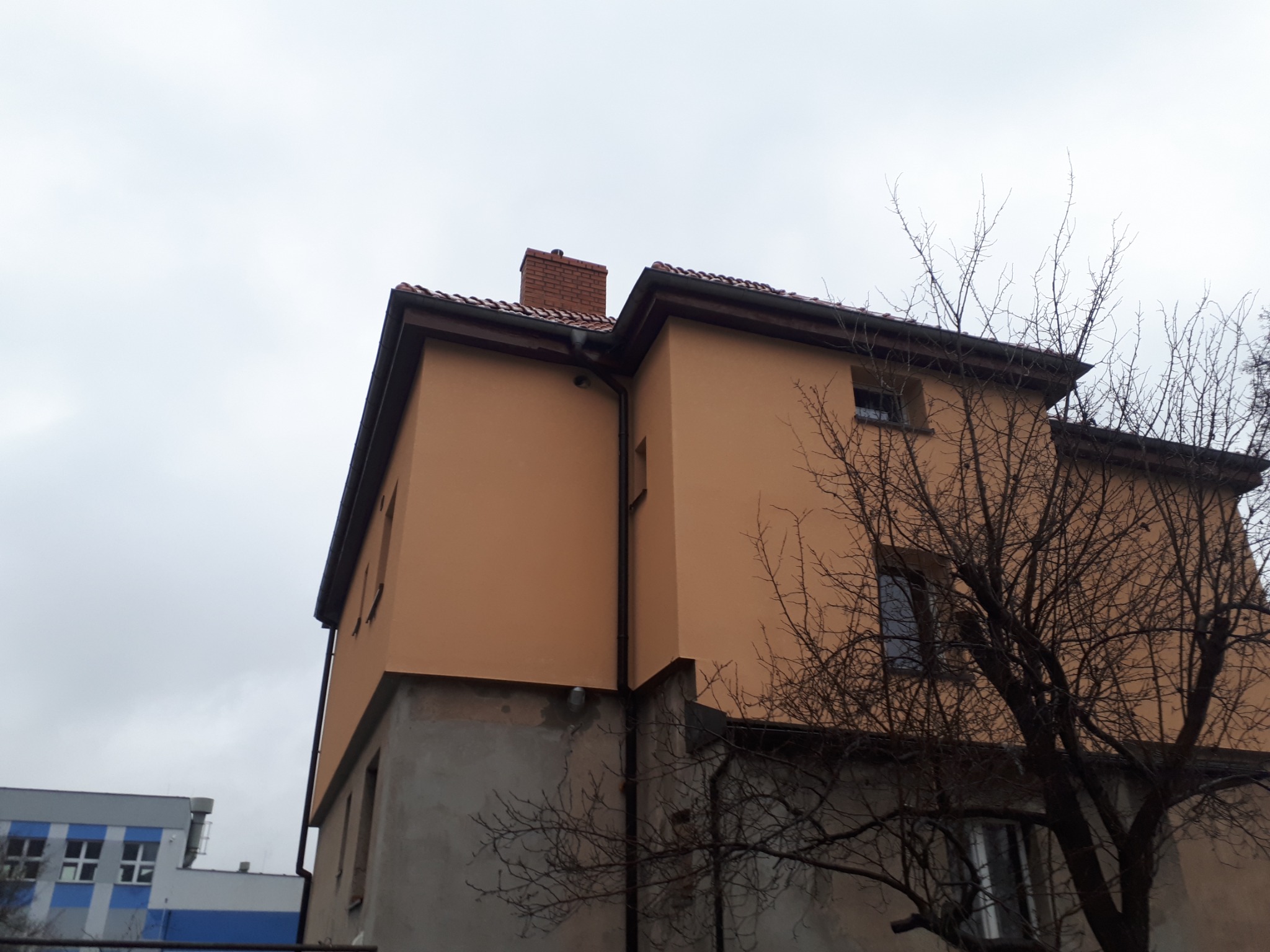 Dwupiętrowy budynek z pomarańczową elewacją i ceglanym kominem widziany z dołu, z widocznymi rynnami i częściowo otynkowanym fundamentem, na tle pochmurnego nieba i fragmentu innego budynku.