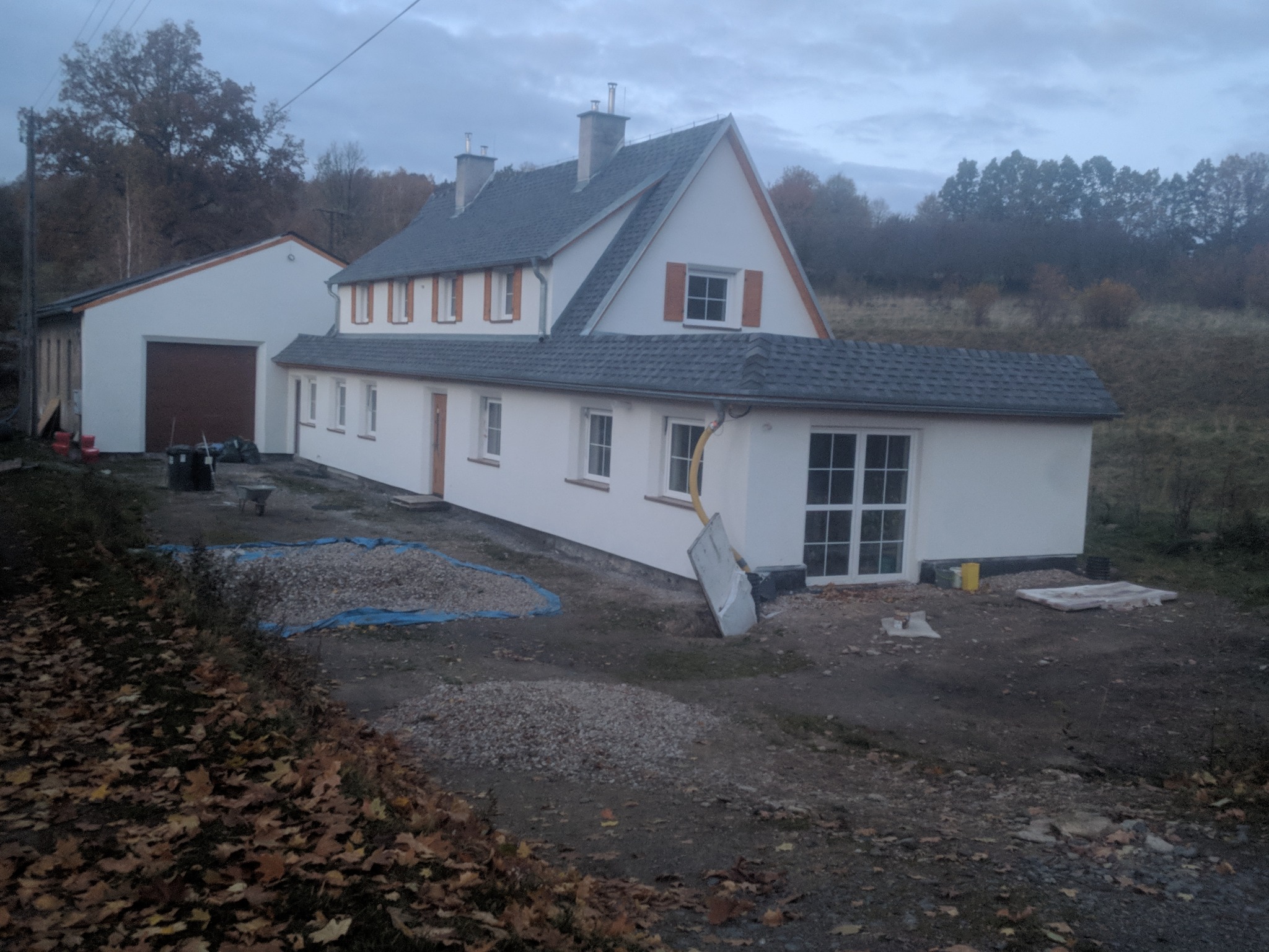 Dom jednorodzinny po remoncie elewacji i dachu, widoczne nowe szare pokrycie dachowe, biała elewacja z drewnianymi okiennicami, przygotowany teren wokół budynku, pochmurne niebo.