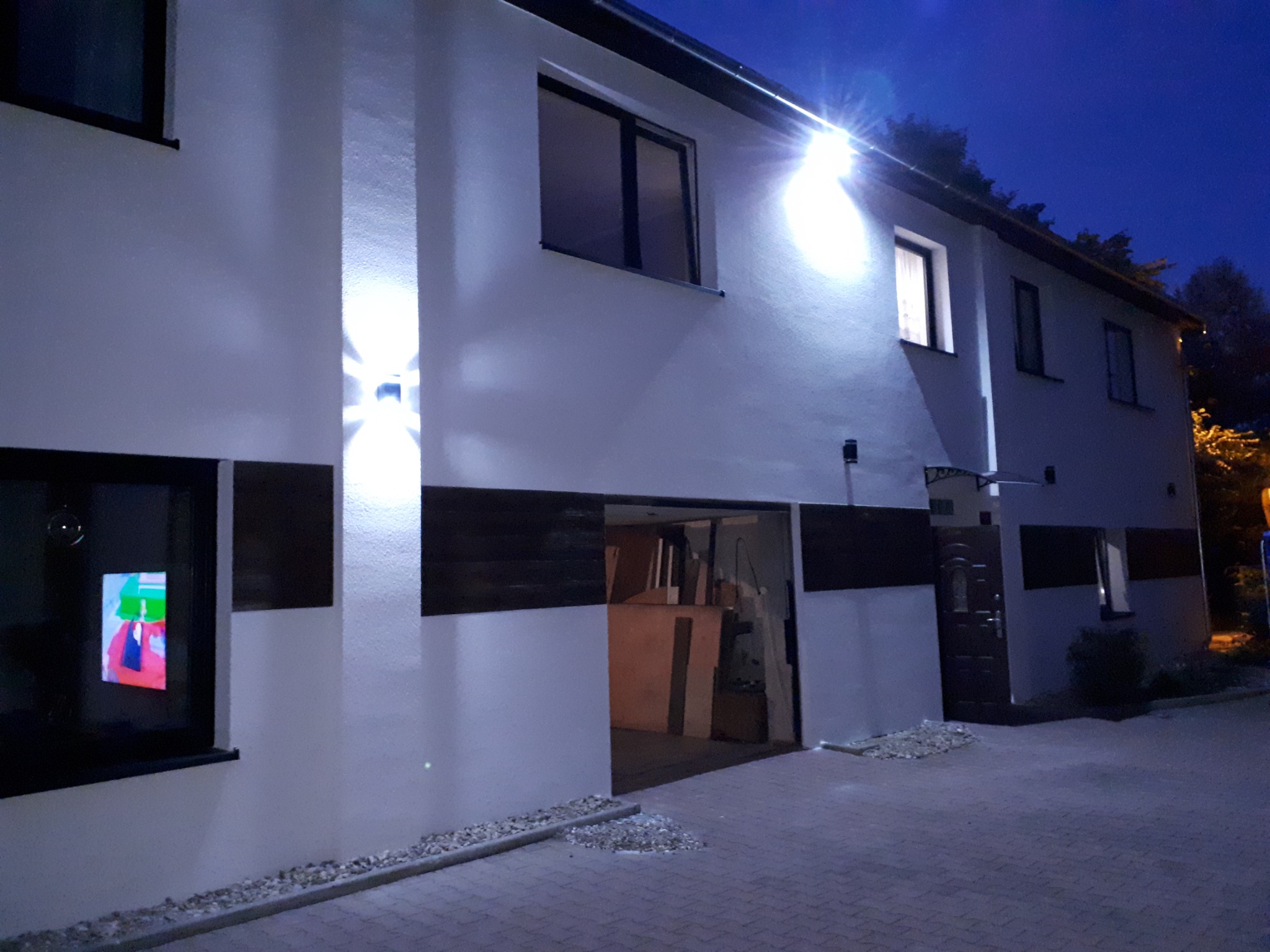 Ocieplona elewacja budynku dwukondygnacyjnego z oknami, oświetleniem zewnętrznym i otwartym garażem, sfotografowana nocą.