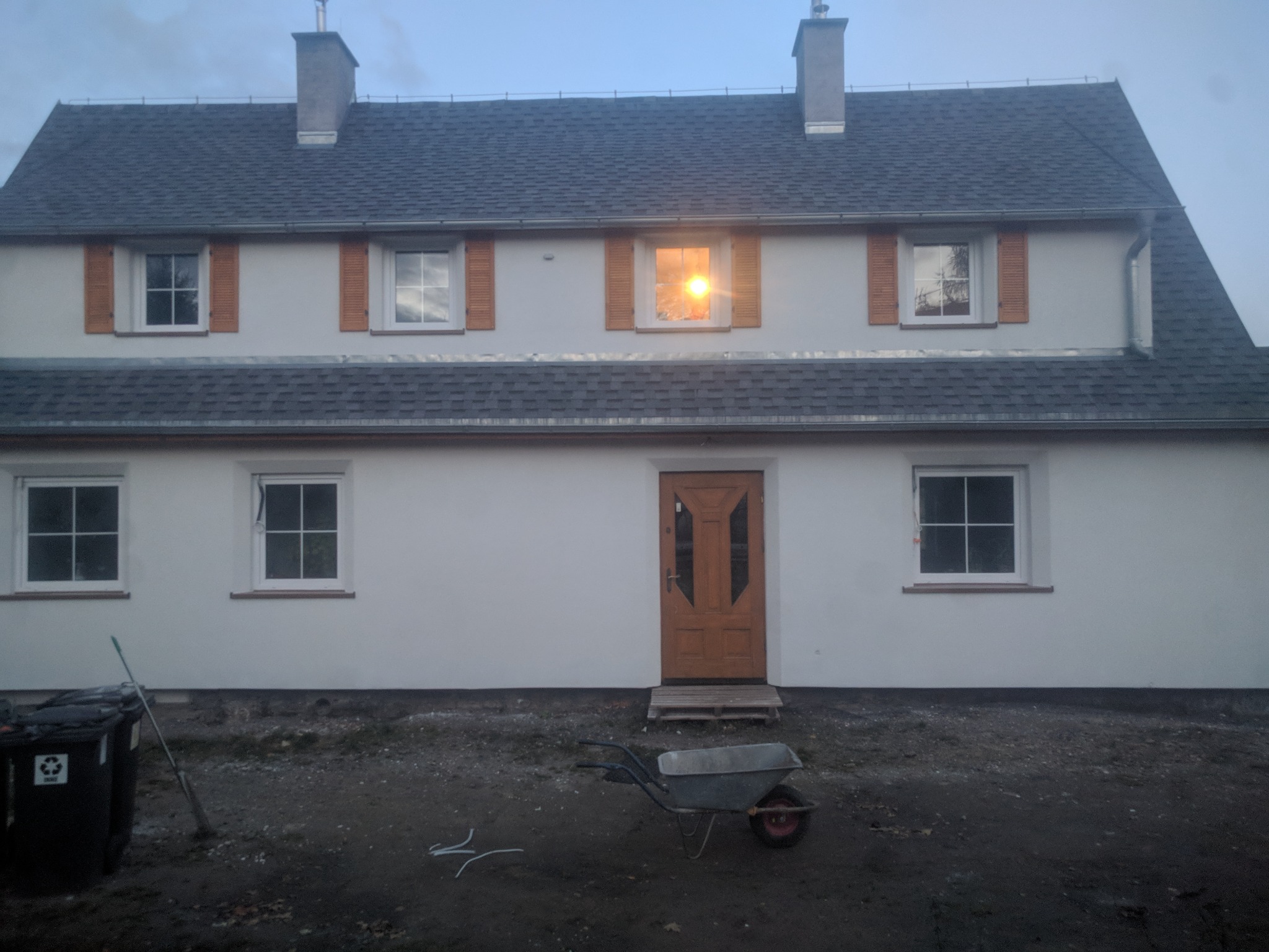 Dwupiętrowy dom z szarą dachówką bitumiczną, drewnianymi okiennicami i świeżo pomalowaną elewacją, widok z zewnątrz, taczka i pojemniki na śmieci na pierwszym planie.