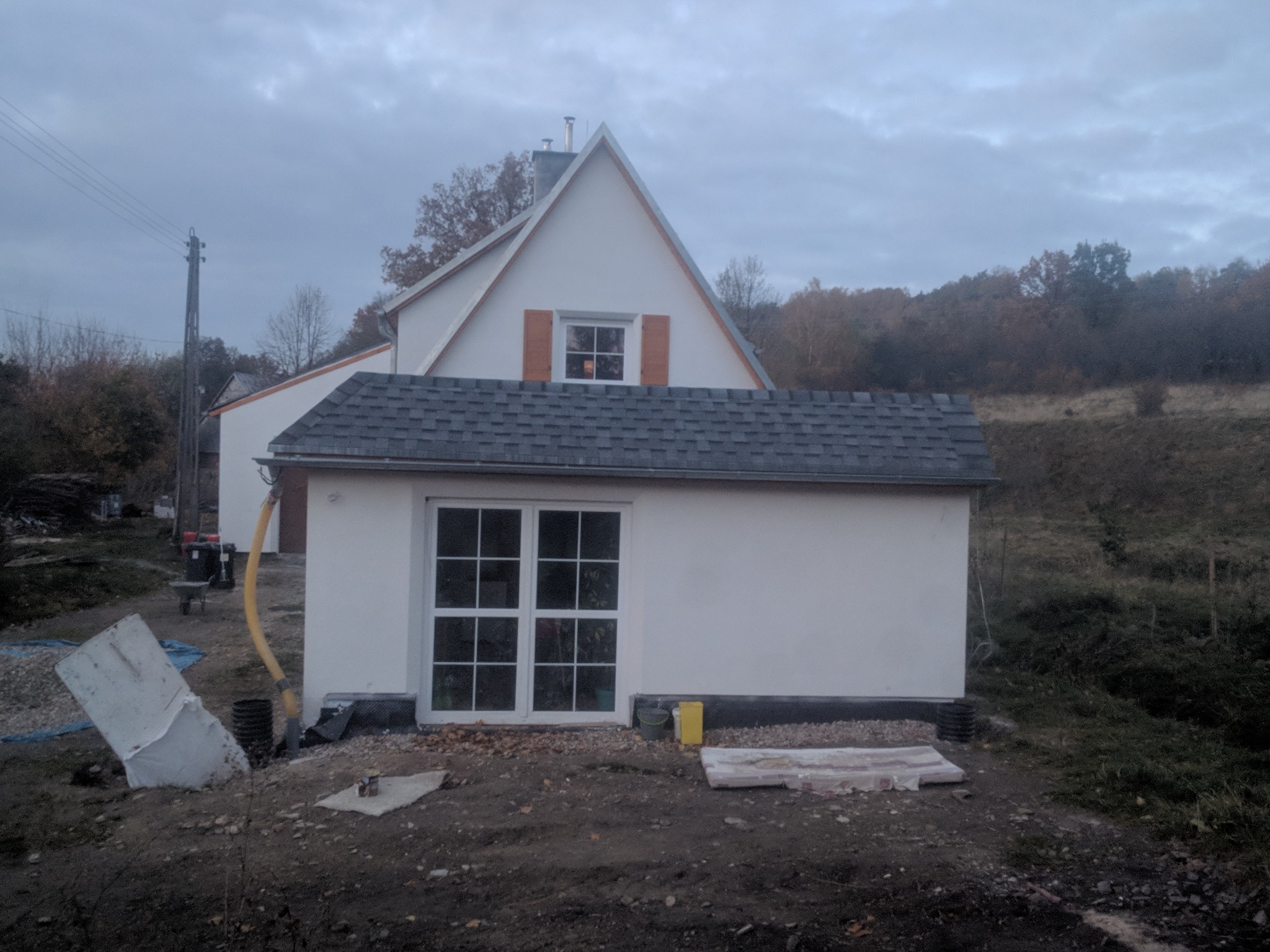 Nowo wykończony, biały budynek z ciemnoszarym dachem z gontów bitumicznych w trakcie prac wykończeniowych, widoczny kabel wentylacyjny i gruz na ziemi.