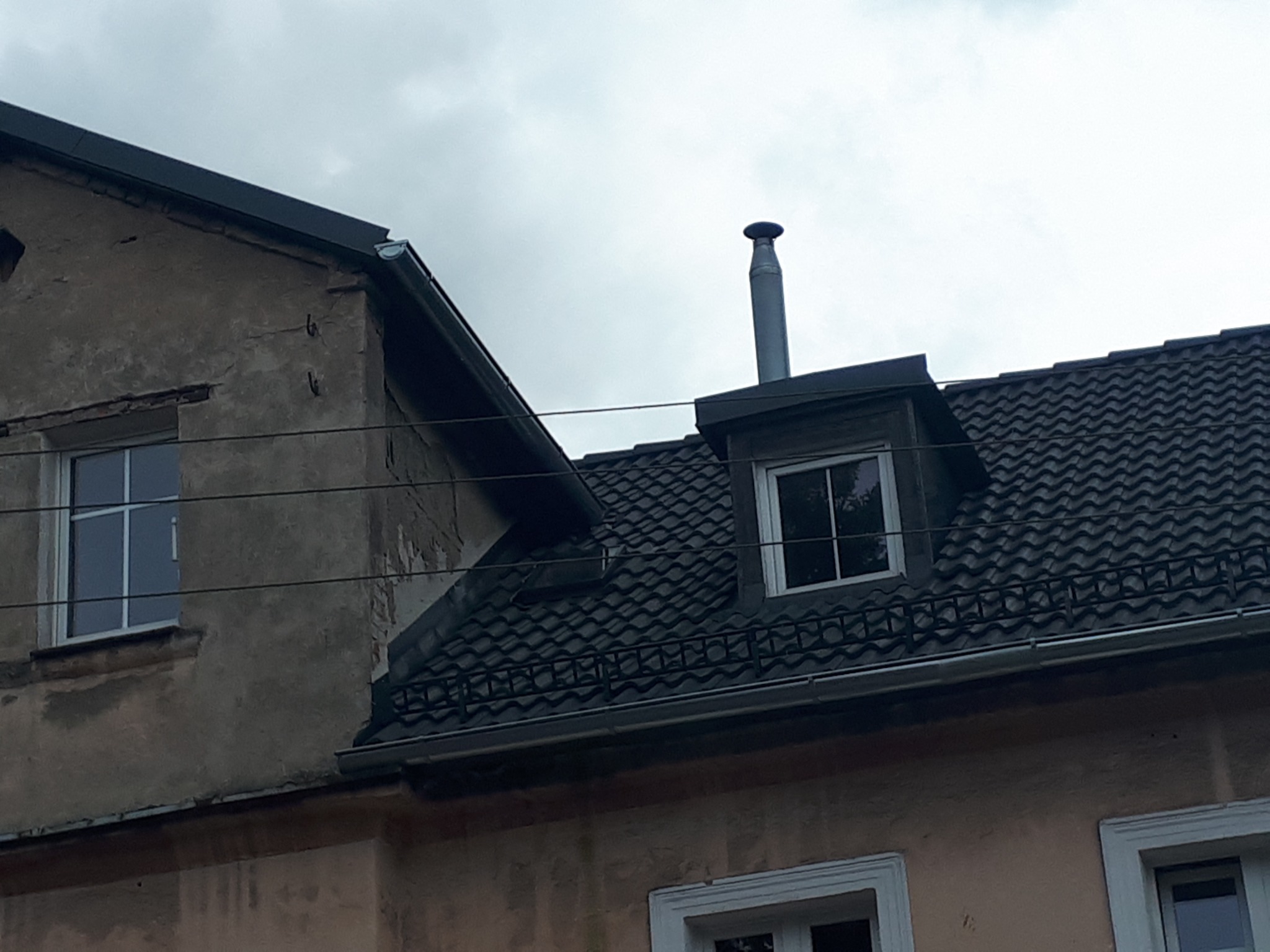 Fragment dachu z czarną dachówką karpiówką, okno dachowe z kominem, widoczne okna w elewacji budynku