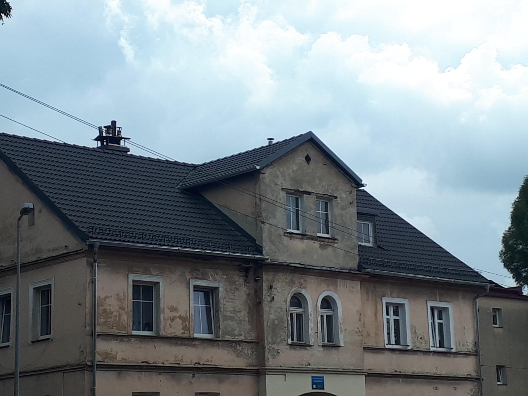 Dwupiętrowy budynek z ciemnym dachem i widocznymi śladami zużycia na elewacji, z elementami architektonicznymi takimi jak okna łukowe i komin na dachu.