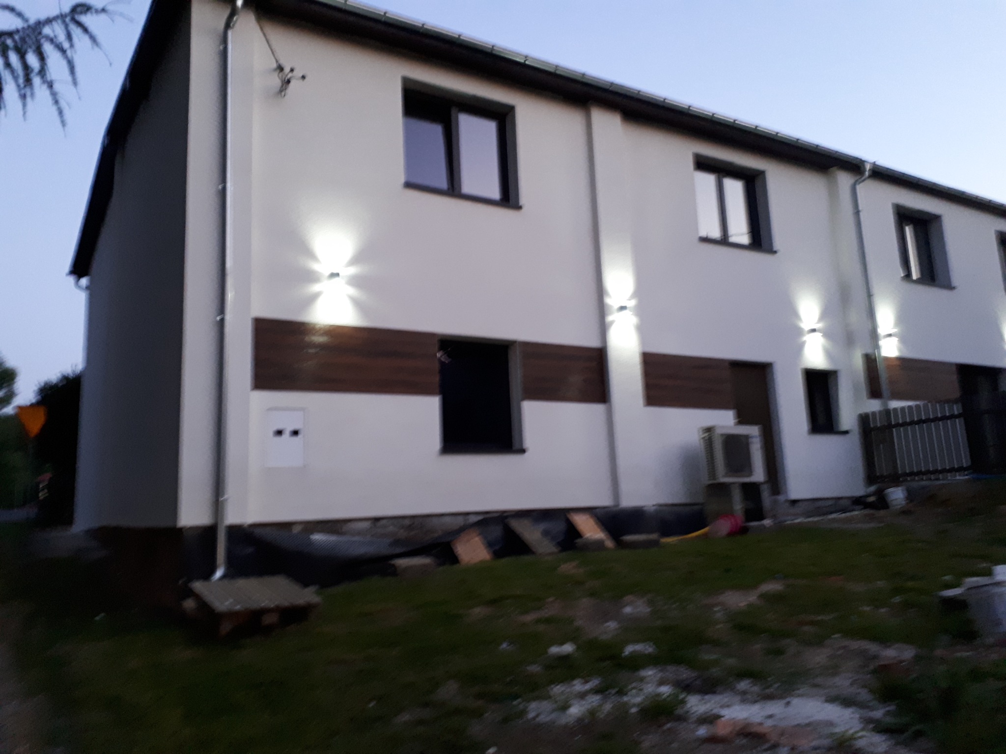 Elewacja szeregowca z oświetleniem zewnętrznym, fragmentem rynny i elementami przygotowanymi do prac ziemnych wokół budynku.