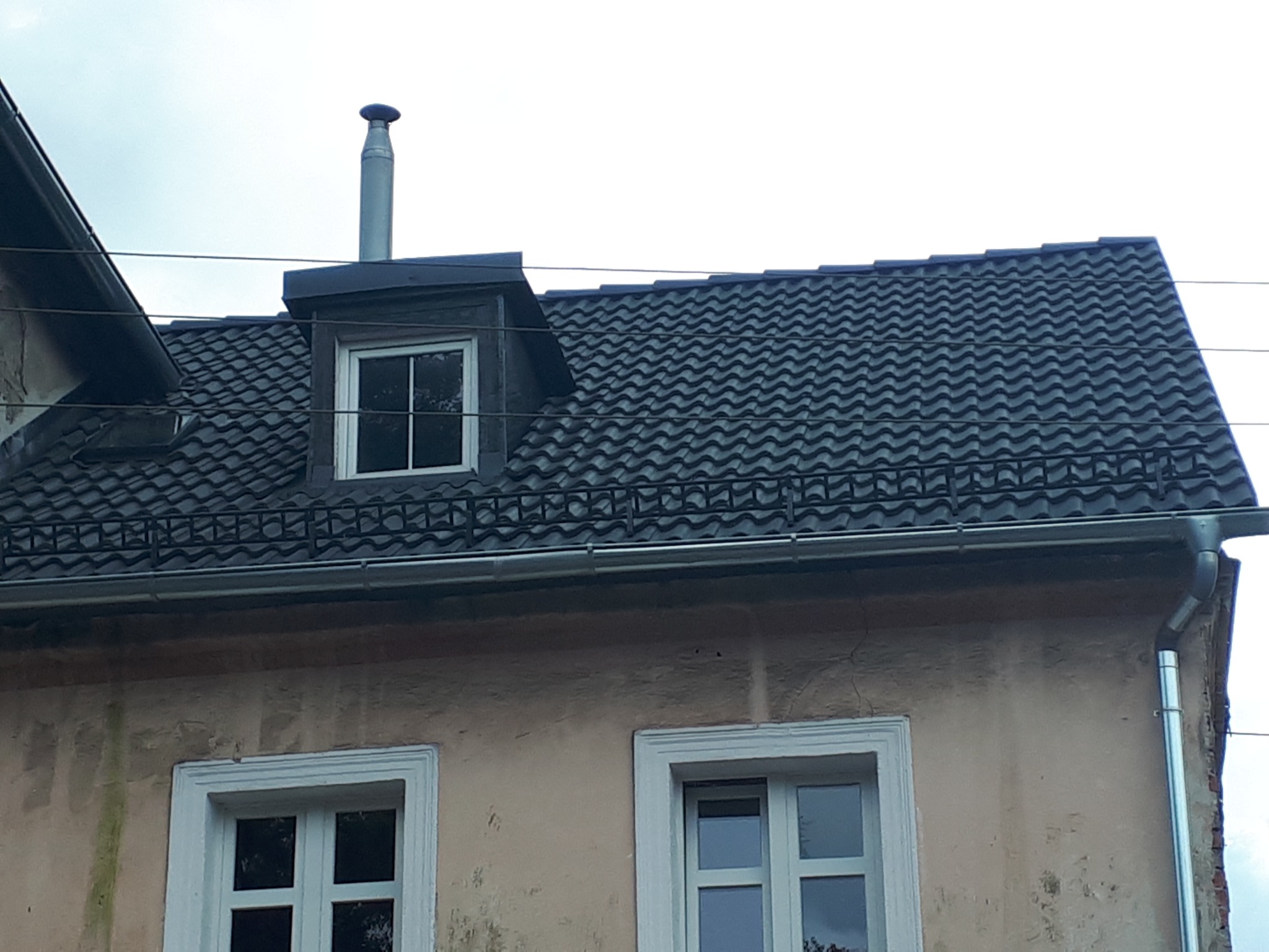 Ciemna dachówka ceramiczna na dachu domu z lukarną i kominem, widoczne rynny i ściana z oknami.