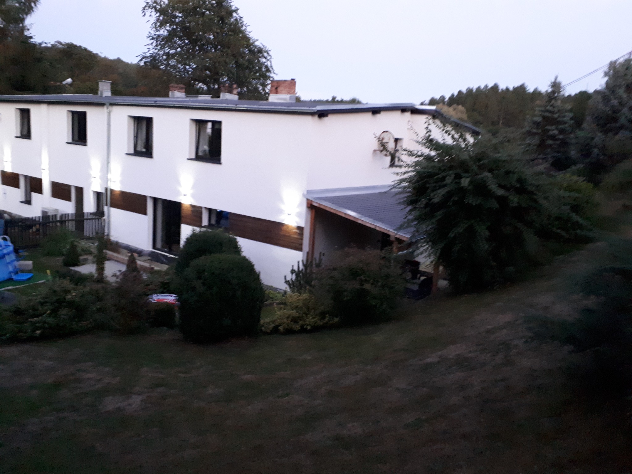 Biały budynek z płaskim dachem i drewnianymi panelami, oświetlony zewnętrznymi lampami, widoczny z perspektywy ogrodu w pochmurny dzień.