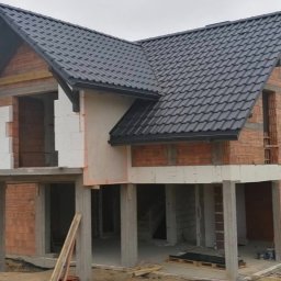 Daniel Tobojko Usługi stolarsko-dekarskie - Dom w trakcie budowy z ciemną dachówką, widoczne cegły, białe bloczki i betonowe podpory, teren budowy.