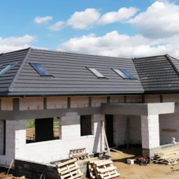 Daniel Tobojko Usługi stolarsko-dekarskie - Dom w trakcie budowy z szarym dachem i oknami dachowymi, widoczne bloczki betonowe i drewniane palety.