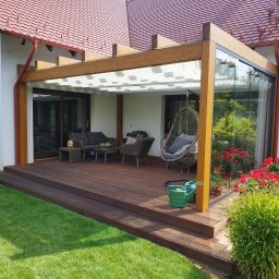 Daniel Tobojko Usługi stolarsko-dekarskie - Zadaszony taras z drewna w Brodnicy, z meblami ogrodowymi i wiszącym fotelem, otoczony zielonym trawnikiem i rabatami kwiatowymi, przylegający do budynku z czerwoną dachówką.