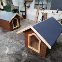 Daniel Tobojko Usługi stolarsko-dekarskie - Dwie brązowe budy dla psa z szarymi dachami, wykonane z drewna, stoją w warsztacie z betonową podłogą i narzędziami w tle.