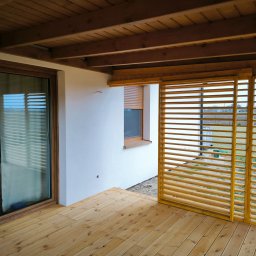 Daniel Tobojko Usługi stolarsko-dekarskie - Zadaszony taras z drewnianą podłogą i przesuwnymi panelami lamelowymi. Widoczne przesuwne drzwi tarasowe i drewniany sufit z belek. Nowoczesne wykończenie.