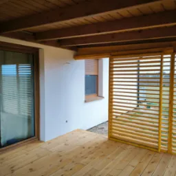 Zadaszony taras z drewnianą podłogą i przesuwnymi panelami lamelowymi. Widoczne przesuwne drzwi tarasowe i drewniany sufit z belek. Nowoczesne wykończenie.