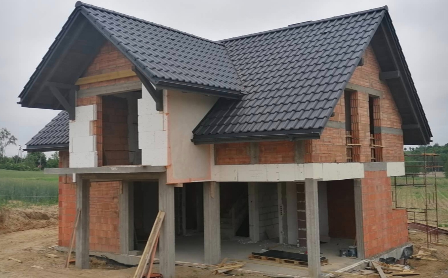 Dom w trakcie budowy z ciemną dachówką, widoczne cegły, białe bloczki i betonowe podpory, teren budowy.