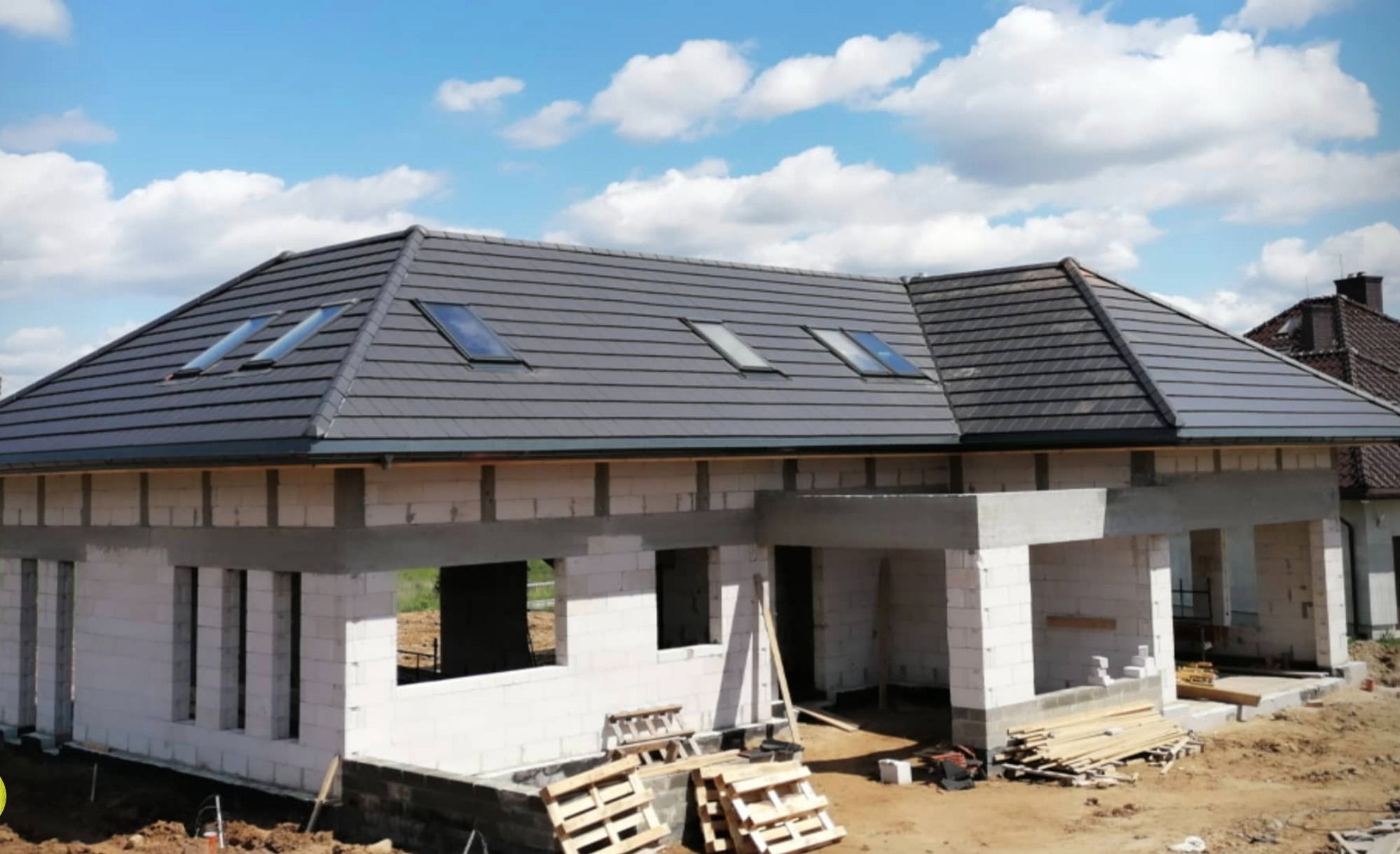 Dom w trakcie budowy z szarym dachem i oknami dachowymi, widoczne bloczki betonowe i drewniane palety.