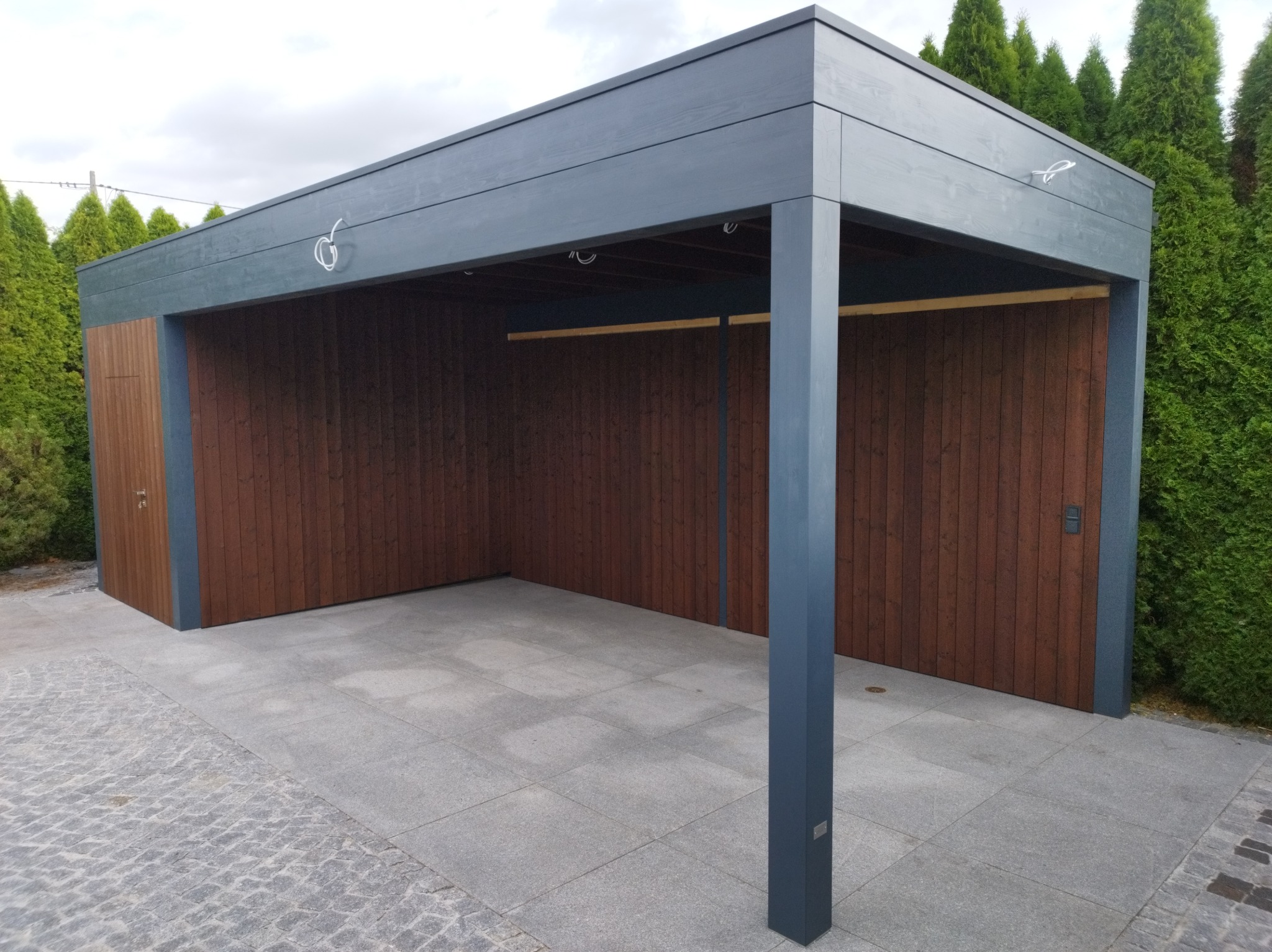 Nowoczesna, drewniana wiata garażowa z płaskim dachem w kolorze grafitowym, zamykana z dwóch stron drewnianymi drzwiami, otoczona tujami i wyłożona kamienną kostką.