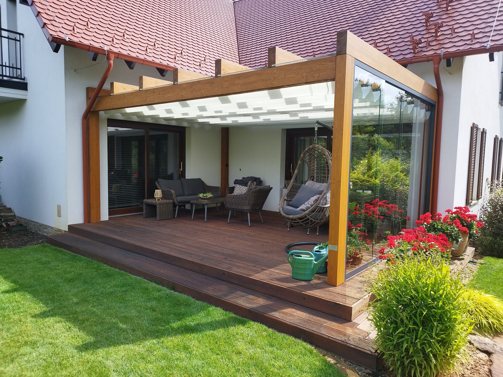 Zadaszony taras z drewna w Brodnicy, z meblami ogrodowymi i wiszącym fotelem, otoczony zielonym trawnikiem i rabatami kwiatowymi, przylegający do budynku z czerwoną dachówką.