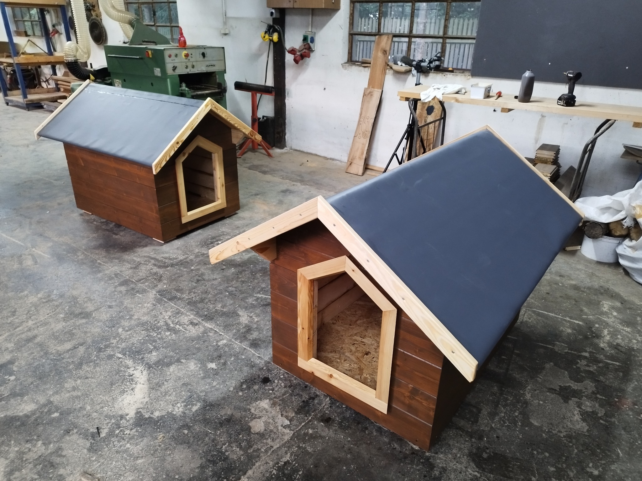 Dwie brązowe budy dla psa z szarymi dachami, wykonane z drewna, stoją w warsztacie z betonową podłogą i narzędziami w tle.