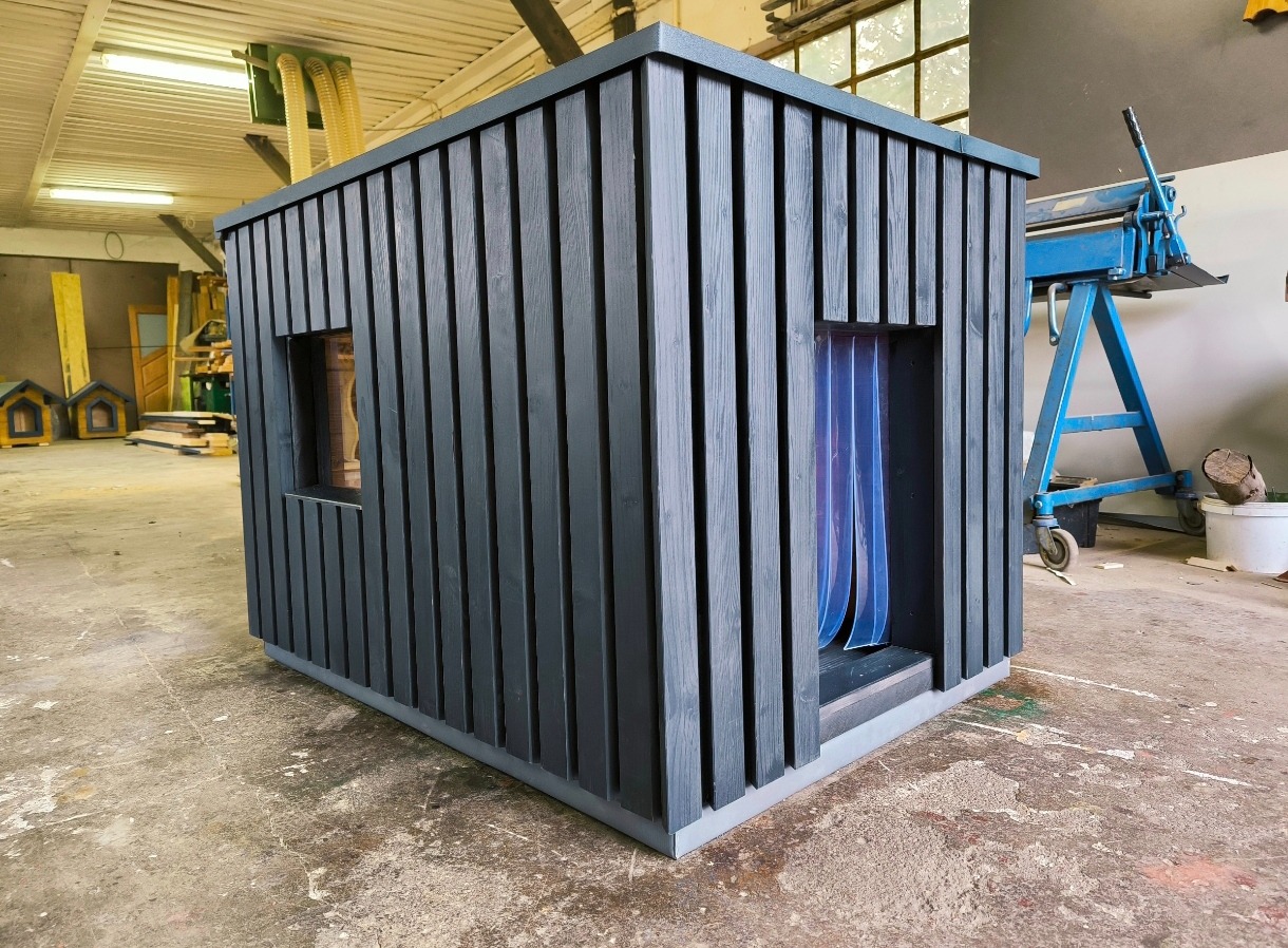 Drewniana konstrukcja w ciemnoszarym kolorze z pionowymi deskami i dwoma otworami, prawdopodobnie buda dla psa lub mała altana, sfotografowana w warsztacie z widocznym sprzętem i innymi...