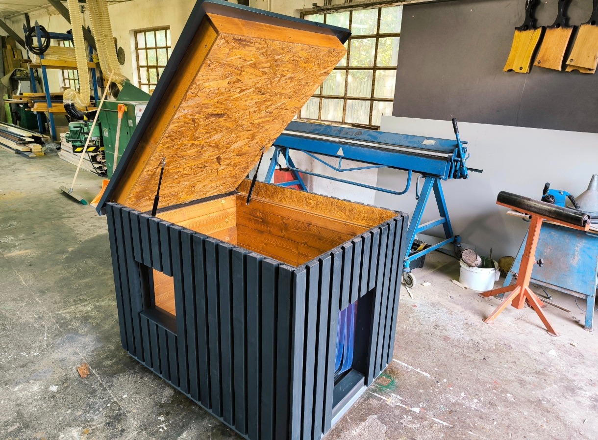 Drewniana konstrukcja z otwieranym wiekiem, pomalowana na ciemny kolor z widocznymi szczelinami, wnętrze wykończone jasnym drewnem, w tle warsztat z narzędziami i maszynami.