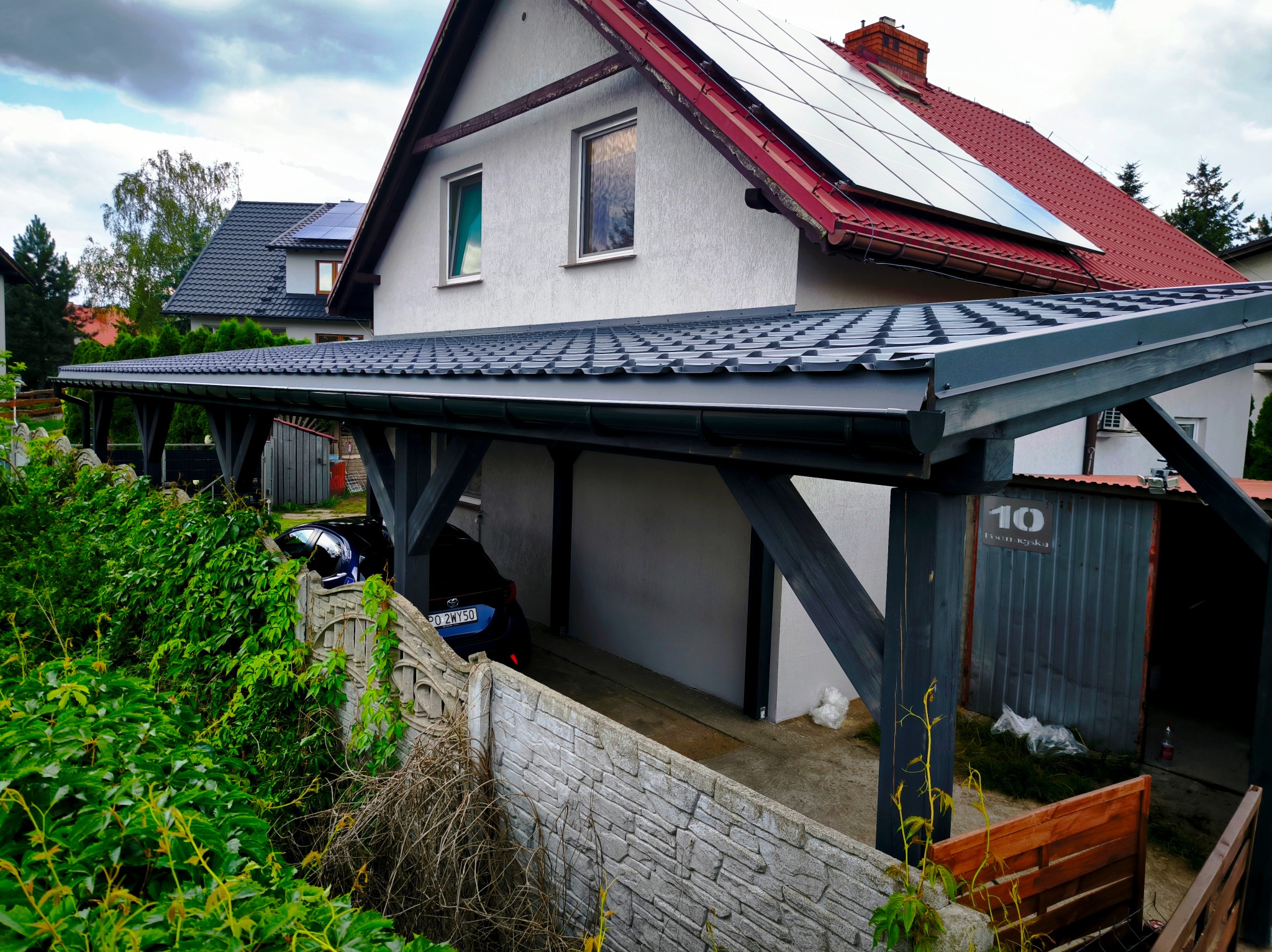 Carport z szarej blachy trapezowej, wsparty na drewnianych słupach, częściowo przysłonięty zielenią. Na dachu domu widoczne panele słoneczne i czerwona dachówka.