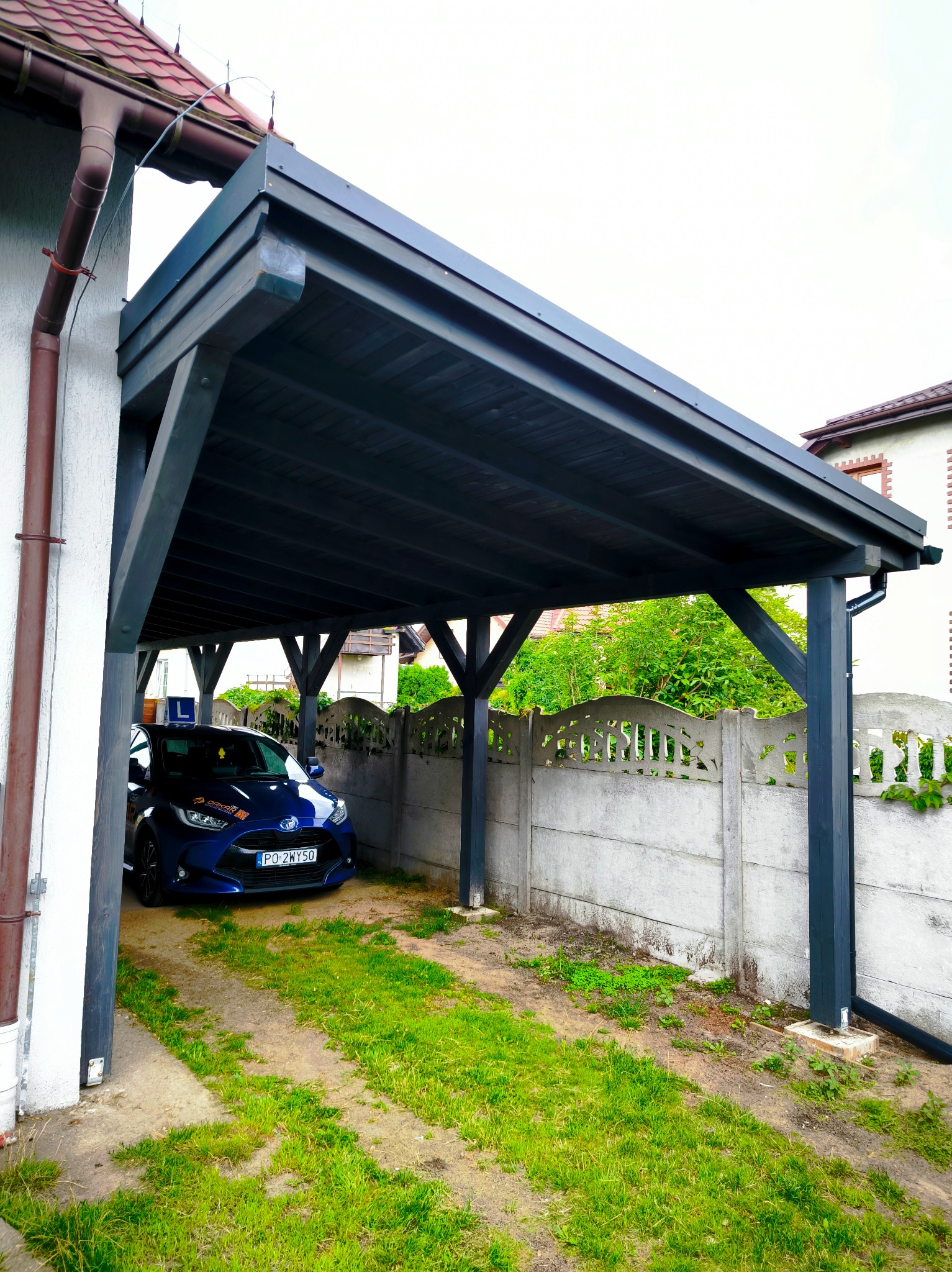 Ciemna, drewniana wiata garażowa przylegająca do budynku z zaparkowanym niebieskim samochodem osobowym. Widoczne elementy konstrukcyjne i fragment posesji.