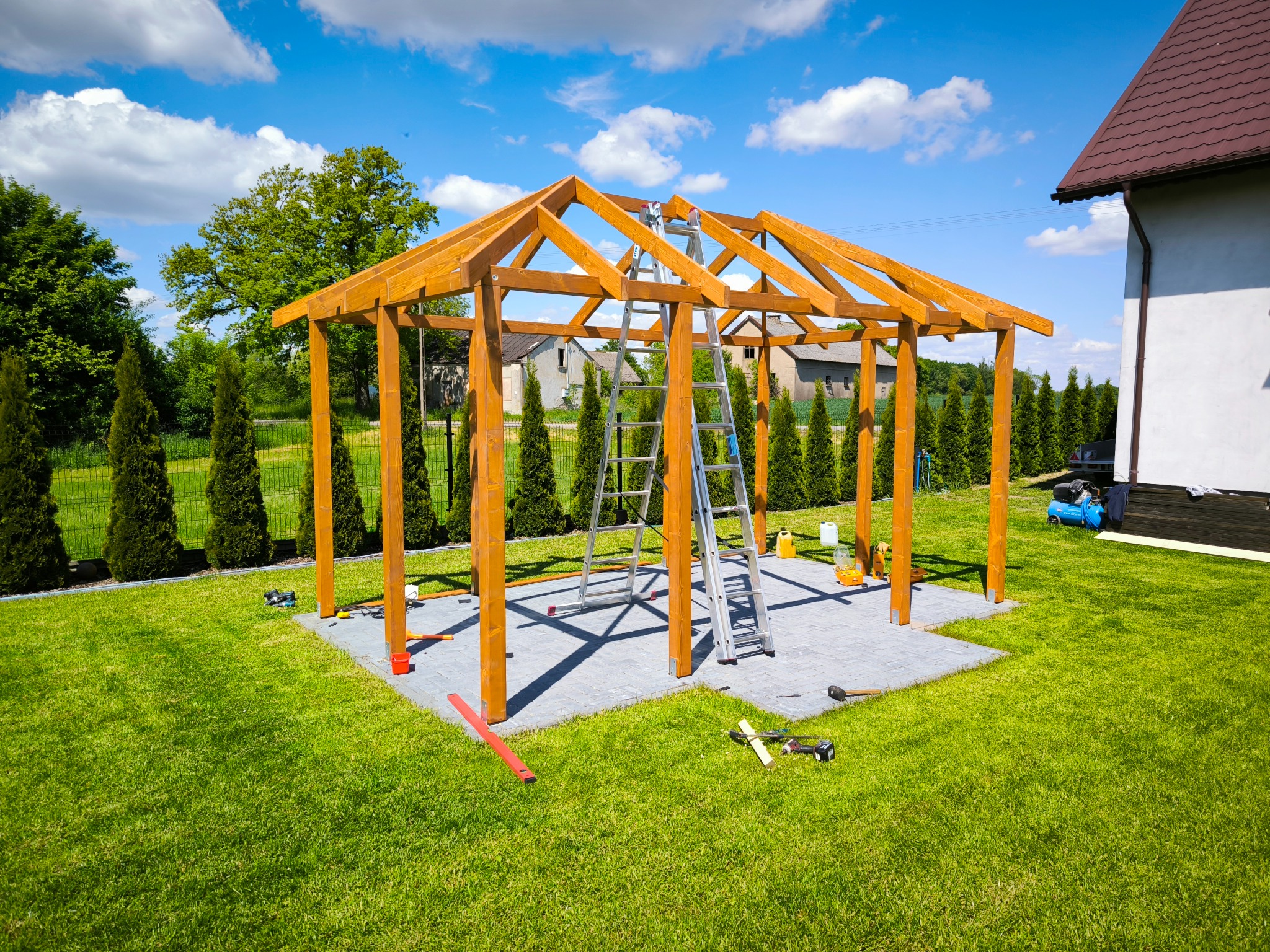Drewniana konstrukcja altany w trakcie budowy, widoczna drabina i narzędzia na kostce brukowej, trawnik, iglaki, dom w tle, słoneczny dzień.