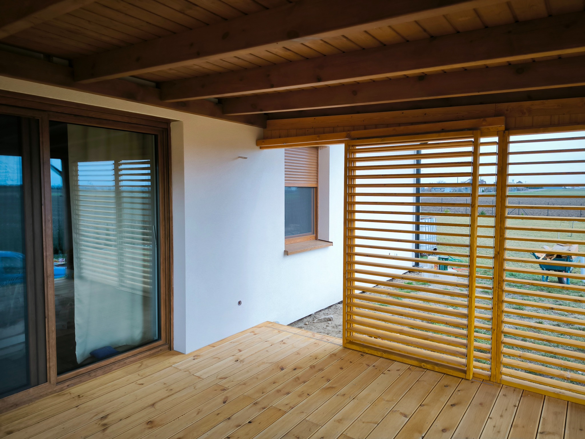 Zadaszony taras z drewnianą podłogą i przesuwnymi panelami lamelowymi. Widoczne przesuwne drzwi tarasowe i drewniany sufit z belek. Nowoczesne wykończenie.