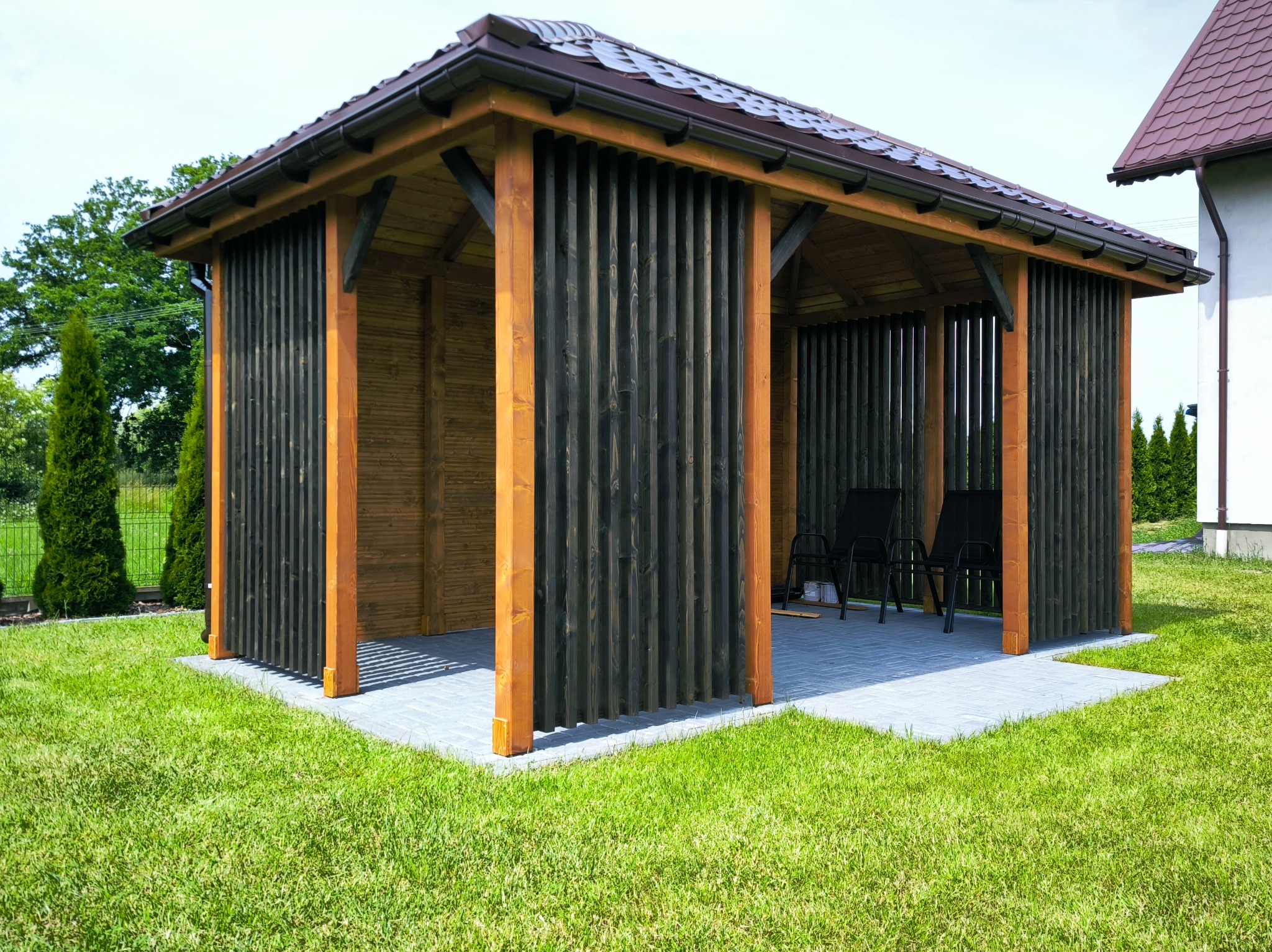 Nowoczesna, drewniana altana z ciemnymi, pionowymi panelami i brązowym dachem na tle zielonego trawnika i budynku. Widoczne krzesła wewnątrz.